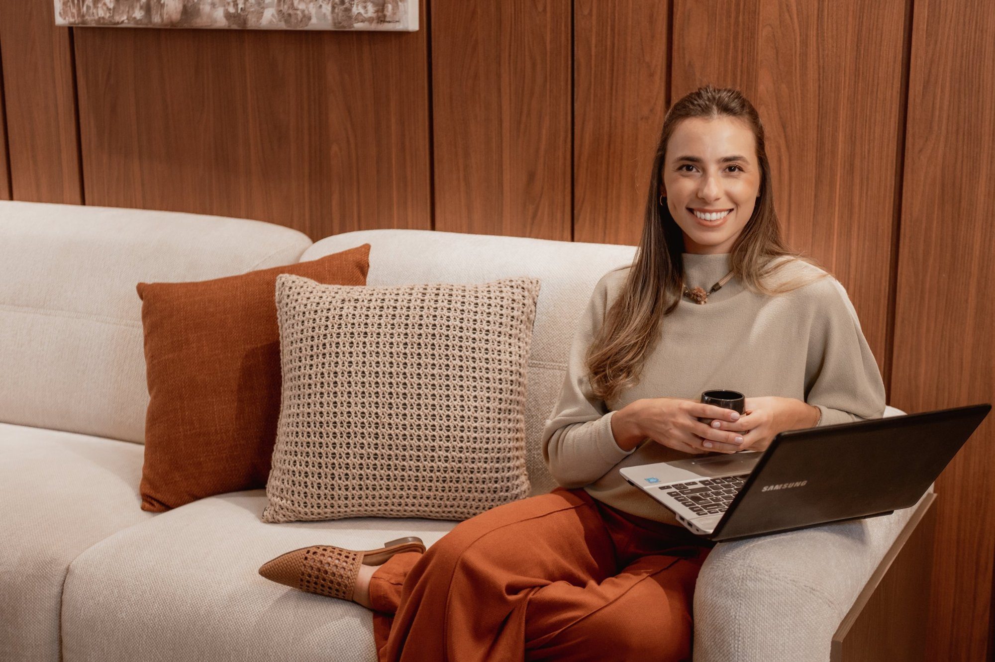 Laís Cohn sorrindo, sentada no sofá com uma xícara e o notebook