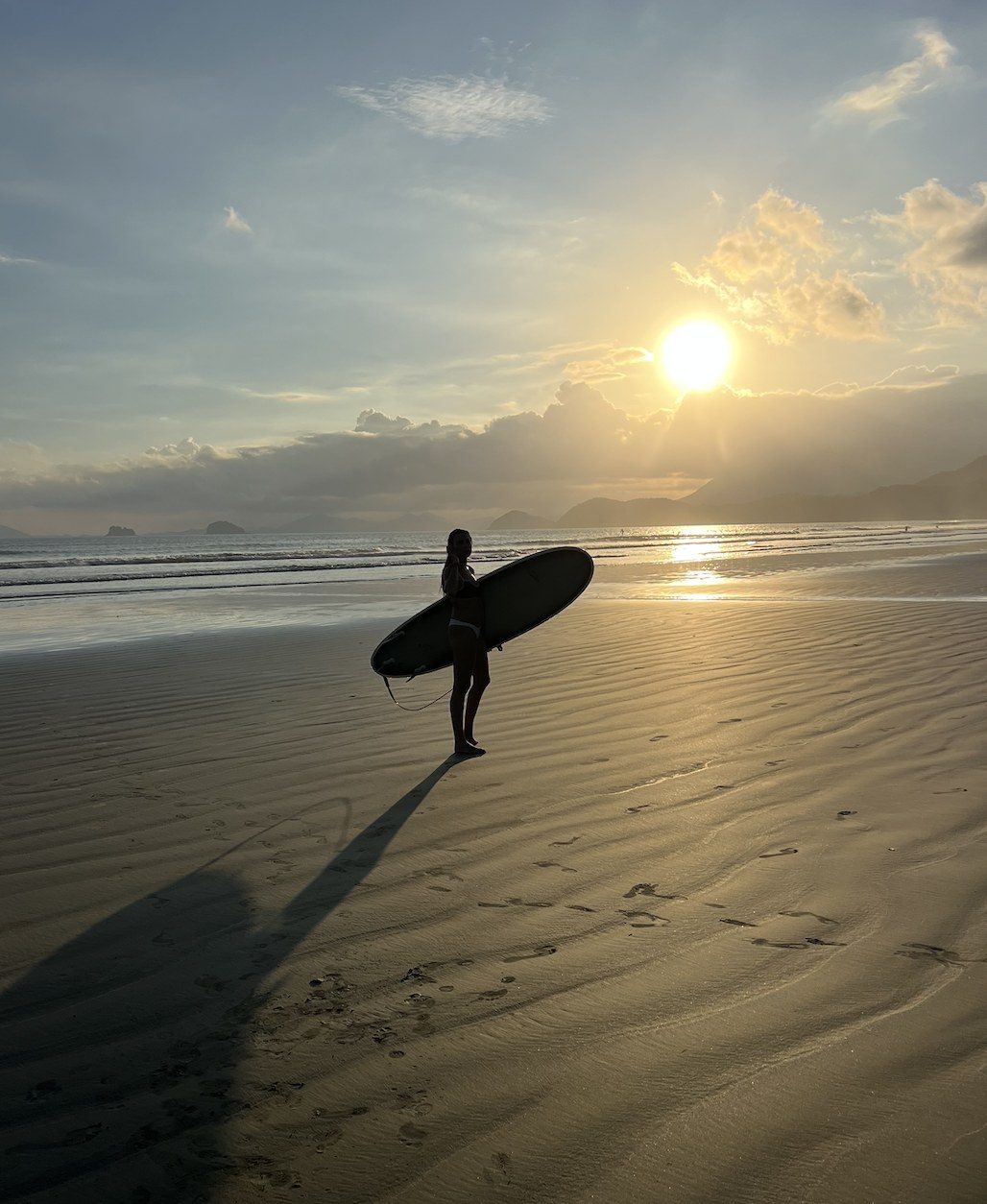 Laís com a prancha de surf na praia ao pôr do sol