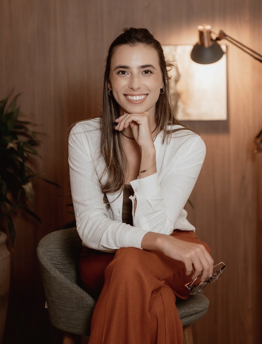 Laís Cohn sorrindo, sentada com a mão no queixo, de camisa branca e calça terracota